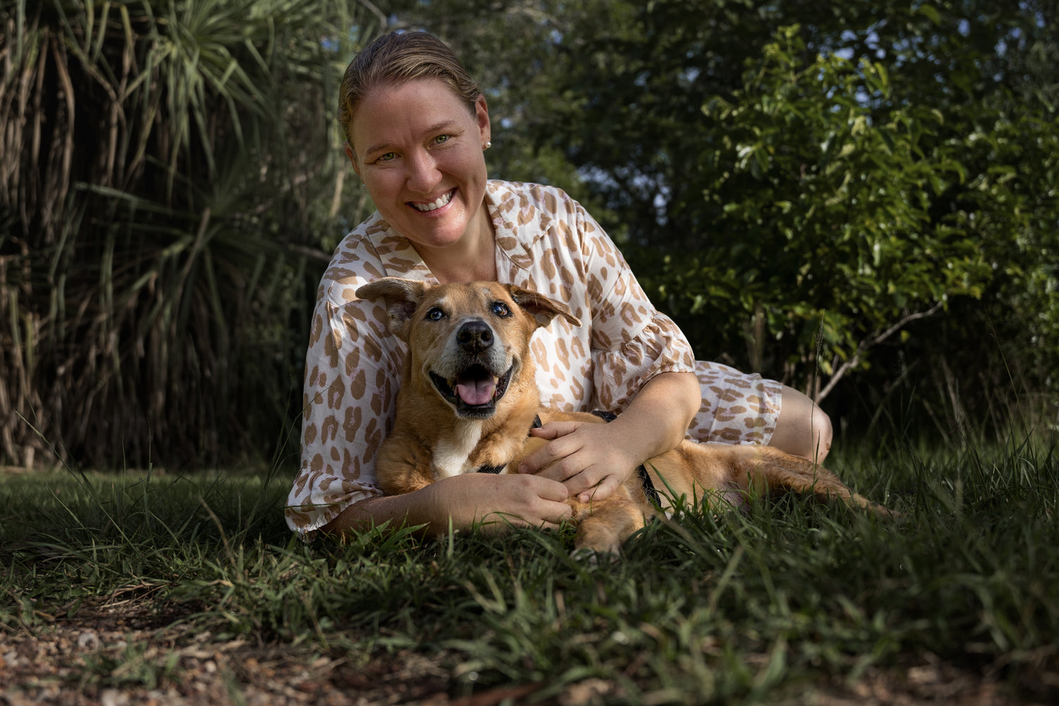 Female in white and brown animal spot dress laying on grass with a tan dog layingin front of her