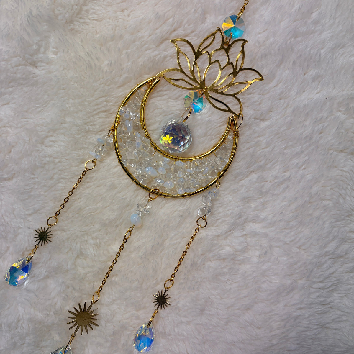 A gold-toned crescent moon and  suncatcher with clear quartz and opolite crystals, displayed on a white faux fur background.