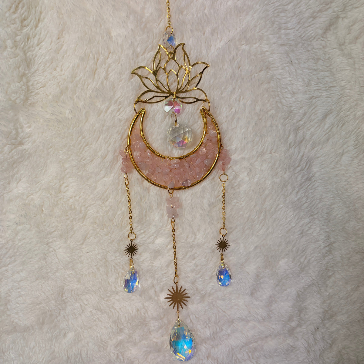 A gold-toned crescent moon and lotus flower hanging sun catcher with rose quartz crystal and iridescent blue rhinestone accents, laid flat on a white faux fur background.