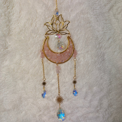 A gold-toned crescent moon and lotus flower hanging sun catcher with rose quartz crystal and iridescent blue rhinestone accents, laid flat on a white faux fur background.