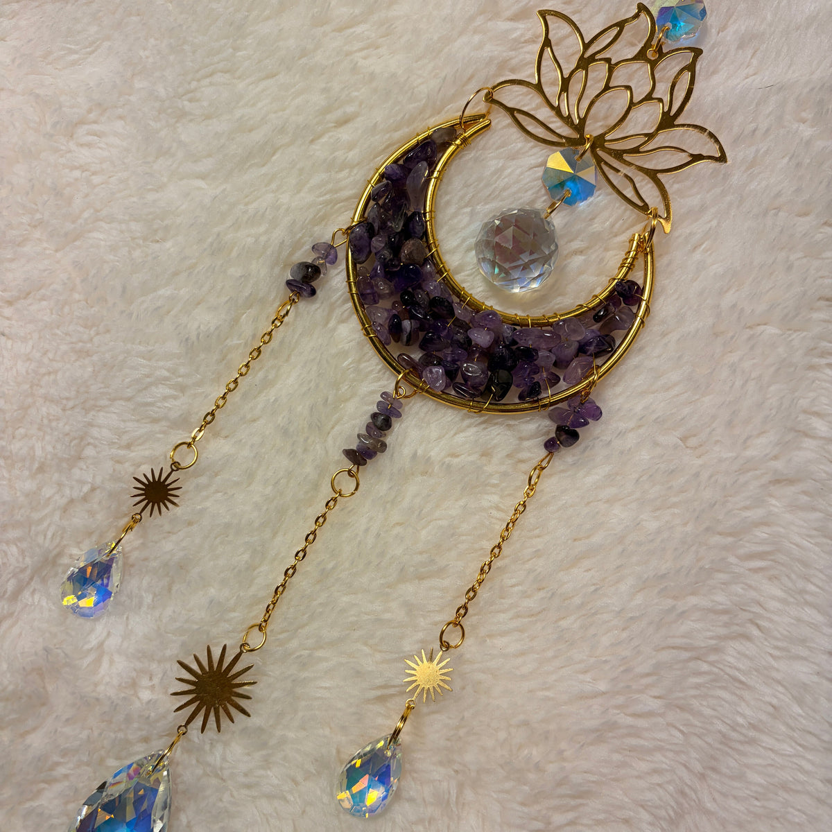 A gold-toned crescent moon and lotus flower hanging sun catcher with  amethyst crystals, starburst sun charms, and faceted crystal drops, displayed on a white faux fur background.