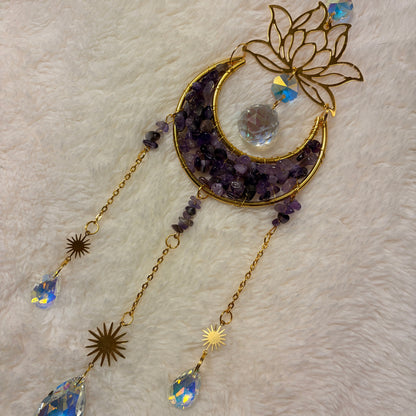 A gold-toned crescent moon and lotus flower hanging sun catcher with  amethyst crystals, starburst sun charms, and faceted crystal drops, displayed on a white faux fur background.