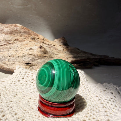 A polished green malachite crystal sphere with swirling bands of dark and light green, displayed on a red circular stand. The sphere is set on a white knitted textile with a piece of driftwood in the background under direct sunlight.