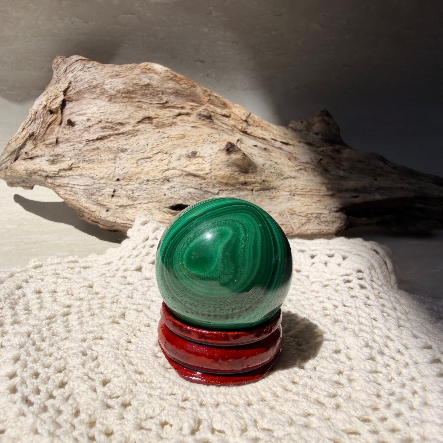 A polished green malachite crystal sphere resting on a wooden stand, placed on a white knitted blanket with a piece of driftwood in the background.
