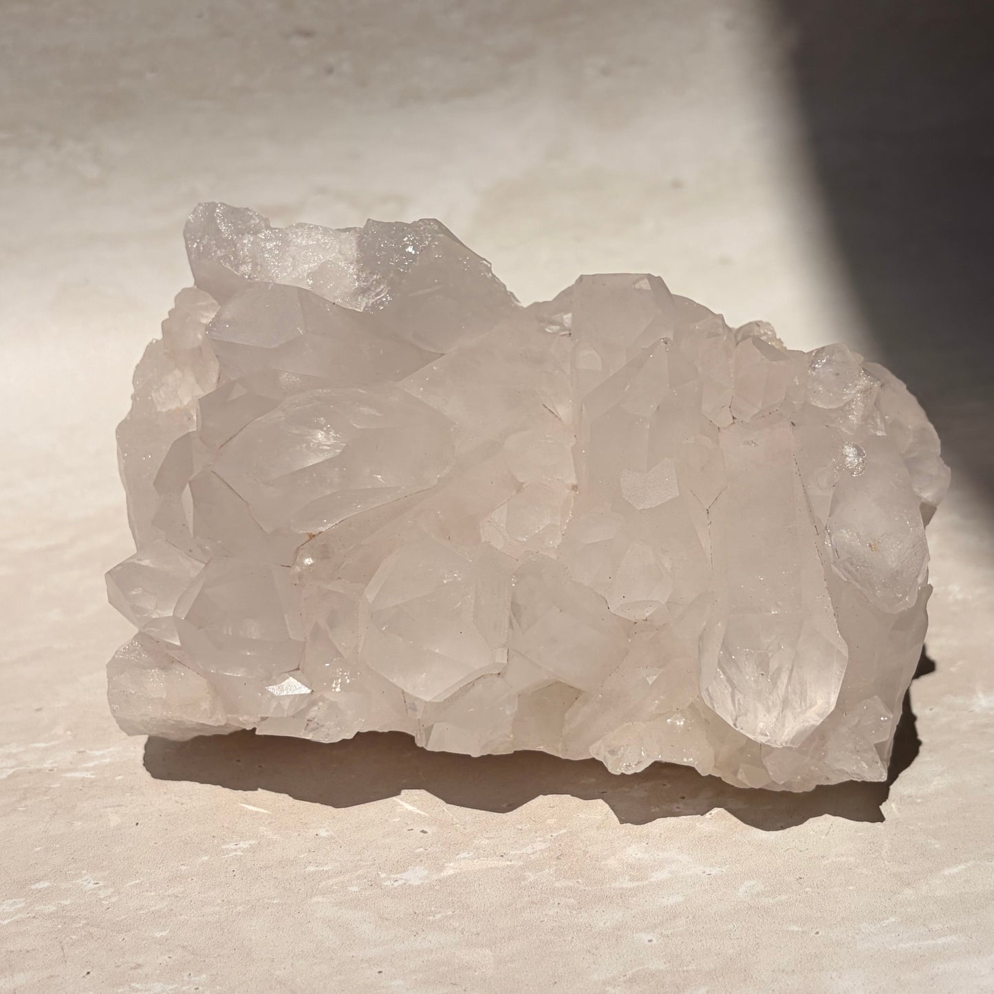 A raw cluster of pale translucent quartz crystals with rough, uneven edges, resting on a light beige stone surface under direct sunlight.