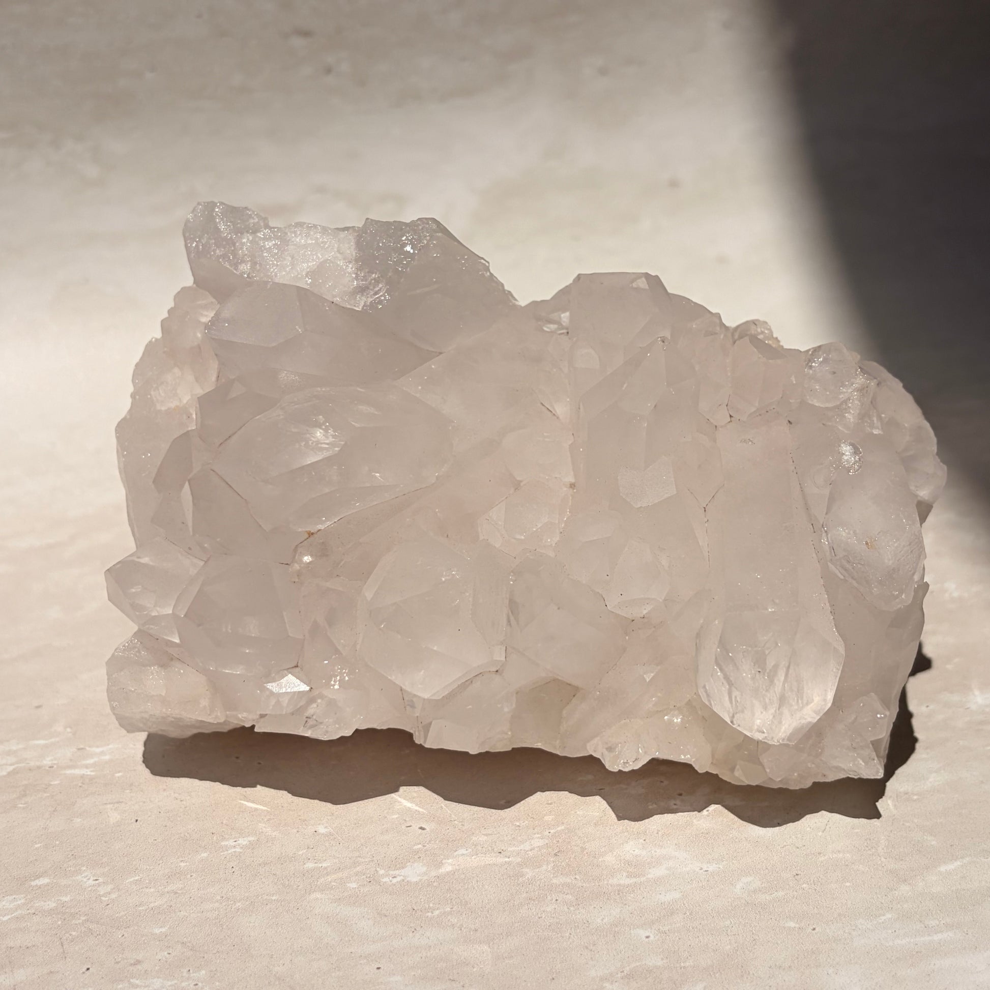 A raw cluster of pale translucent quartz crystals with rough, uneven edges, resting on a light beige stone surface under direct sunlight.
