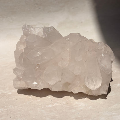 A raw cluster of pale translucent quartz crystals with rough, uneven edges, resting on a light beige stone surface under direct sunlight.