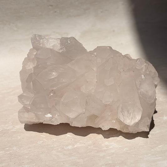 A raw cluster of pale translucent quartz crystals with rough, uneven edges, resting on a light beige stone surface under direct sunlight.